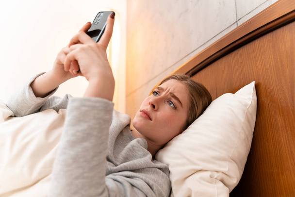 Photo of a sad pretty young woman indoors at home lies in bed using mobile phone. - Fotoğraf, Görsel
