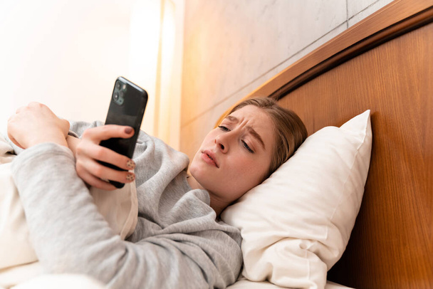 Photo of a sad pretty young woman indoors at home lies in bed using mobile phone. - Photo, Image