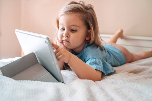Beautiful child little girl playing with a tablet while lying on bed at home.  - Фото, изображение