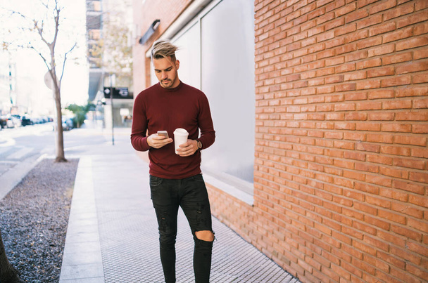 Portrait of young handsome man using his mobile phone while holding a cup of coffee outdoors in the street. Communication concept. - Foto, immagini