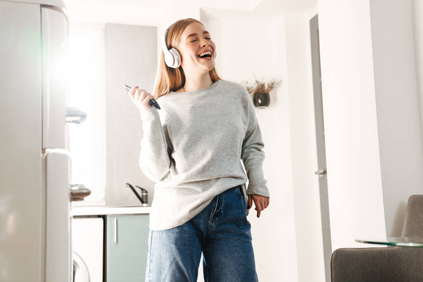 Photo of young cute positive girl holding mobile phone listening music with headphones indoors at home at kitchen. - Foto, imagen