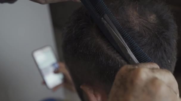 Closeup on woman who cutting her husband hair sitting and looking at smartphone browsing social networks at home during the coronavirus pandemic. Hairdresser concept. Prores 422 - Кадри, відео