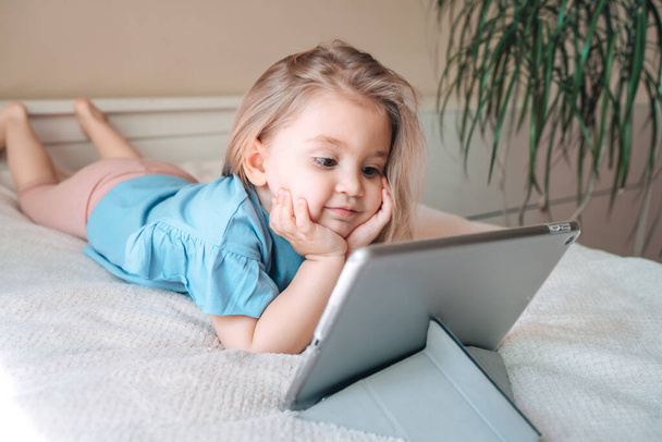 Beautiful child little girl playing with a tablet while lying on bed at home.  - Φωτογραφία, εικόνα