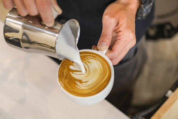 Primer plano de la mano barista verter leche triturada en la taza blanca de café caliente para crear arte de café con leche
. - Foto, Imagen