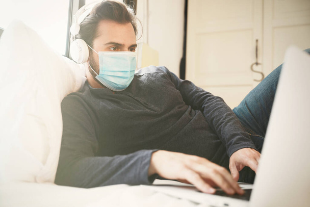 Shot of young man in a medical mask using laptop for work at home. Quarantine coronavirus, pandemia covid19. - 写真・画像