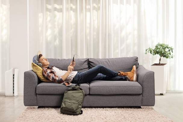 Guy lying on a sofa at home and listening to music on headphones - Foto, Imagen