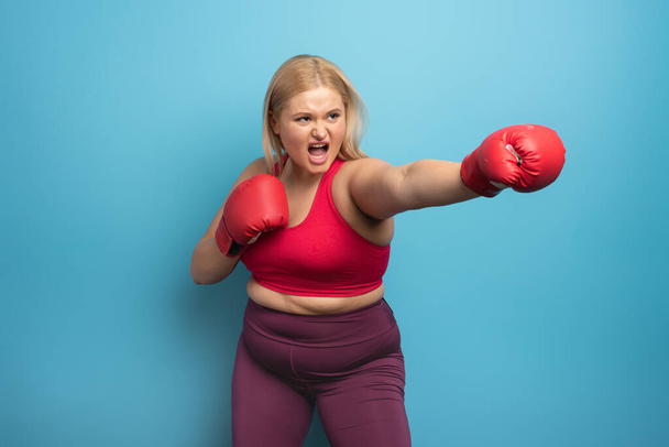 Fat girl in fitness suite does boxing. Cyan background - Фото, зображення