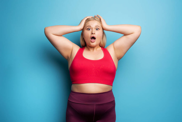 Fat girl is worried because the scale marks a high weight. Cyan background - Φωτογραφία, εικόνα