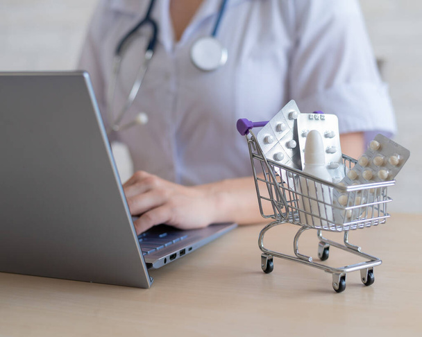 Unrecognizable female doctor prints a report on a laptop. Online pharmacy concept. The faceless pharmacist sells drugs on the Internet. Mini shopping trolley beatery tablets. - Foto, Bild