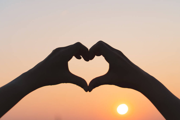 Γυναίκα χέρι κάνει σχήμα καρδιάς στο ηλιοβασίλεμα ουρανό και bokeh φόντο. - Φωτογραφία, εικόνα