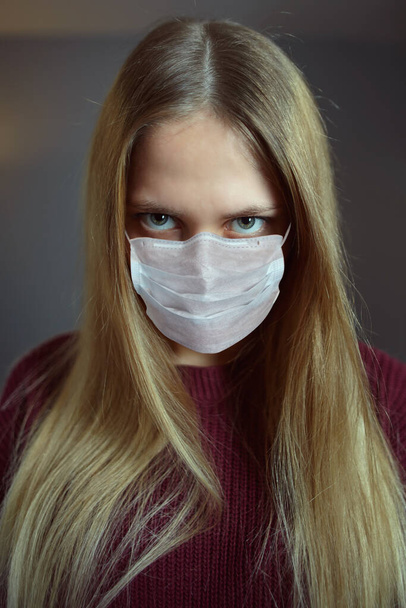 Young Caucasian woman with paranoid problems acquired in home isolation indoors during pandemic of coronavirus infection Covid-19. Crazy look of white female who wears protective face mask. - Foto, afbeelding