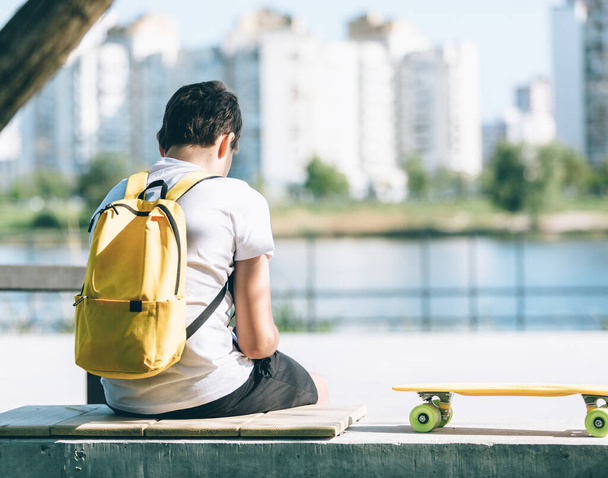 Rückenansicht eines kleinen Jungen mit gelbem Rucksack und Skateboard im Stadtpark. Trendige Studentin sitzt draußen und blickt nach vorn. Jugend, Freizeit, Hobby, Freizeitkonzept - Foto, Bild