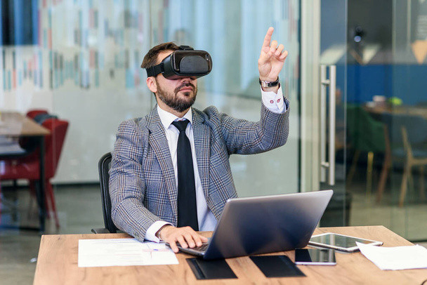 Young Caucasian bearded businessman in blue suit using vr goggles at office. - Fotó, kép