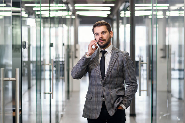 Successful business man have telephone conversation while standing in empty office corridor. Male professional banker in suit talking on mobile phone during work break. - Фото, изображение