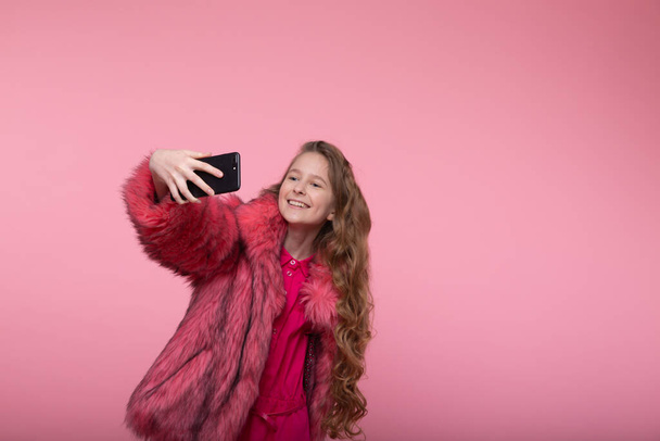 Mooi jong meisje maakt selfie aan de telefoon in modieuze kleren in een bontjas in roze bont en een roze jurk - Foto, afbeelding