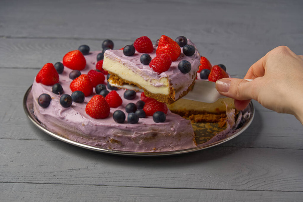 Woman divided cheesecake and take one slice to offer it. Home made Cheesecake with Strawberry, blueberry and raspberry on grey wooden background. - Photo, Image
