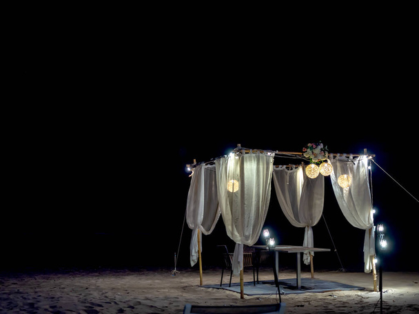 Romantyczny stół, ozdoba z drewna, lampa i biały materiał na plaży w nocy na ciemnym tle. - Zdjęcie, obraz