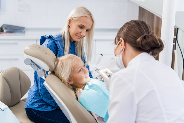 Dişçi kızın dişlerini özel bir aletle temizliyor, kız doktora bakıyor. - Fotoğraf, Görsel