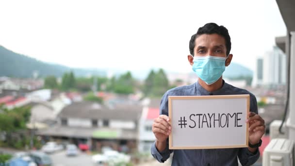 医療用マスクの若いアジア人男性,カメラを見て、図面を表示"#STAYHOME"病気やほこりを防ぐために,午後5時,家庭用検疫コロナウイルスパンデミック予防に滞在.スペースのコピー. - 映像、動画