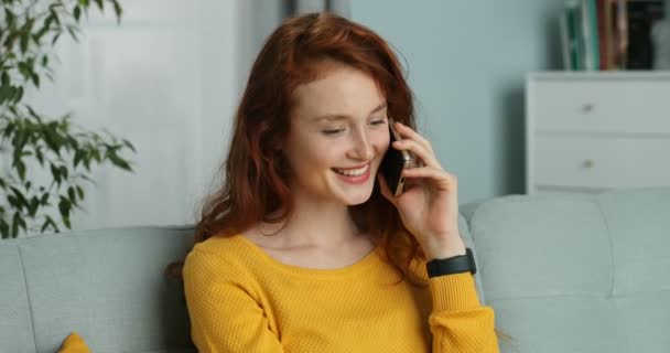 Joyful young Caucasian red-haired woman talking on cellphone and smiling and siting on couch at home. Pretty happy girl with red curly hair speaking on mobile phone in living room. - Video