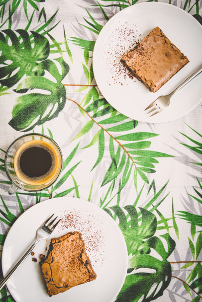 Horní pohled na šálek kávy a dva dezertní talíře s koláčem přes ubrus se zelenými listy. - Fotografie, Obrázek