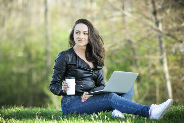 Kahve fincanı ve dizüstü bilgisayarı olan siyah elbiseli genç bir kadın parkta çalışıyor. Uzaktan kumanda. Dijital çalışma. Uzaklık öğrenimi - Fotoğraf, Görsel