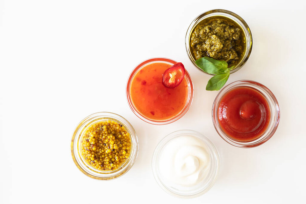Ketchup, sweet chili sauce, pesto, french mustard and mayonnaise in glass bowls on a white background, top view. Various seasoning and dip with copy space - Photo, Image