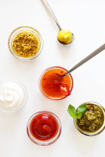 Ketchup, süße Chilisoße, Pesto, französischer Senf und Mayonnaise in Glasschüsseln und Löffeln auf weißem Hintergrund, darüber. Verschiedene Gewürze und Dip, vertikal - Foto, Bild