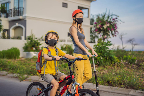 Active school kid boy and his mom in medical mask and safety helmet riding a bike with backpack on sunny day. Happy child biking on way to school. You need to go to school in a mask because of the - Foto, Imagen