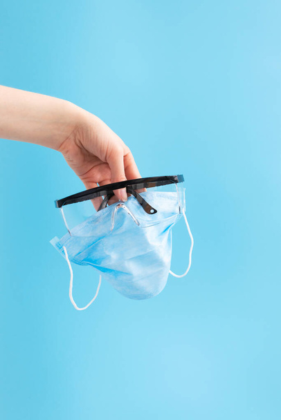 Gafas protectoras de plástico y máscara quirúrgica sanitaria desechable en mano femenina sobre fondo azul
 - Foto, Imagen