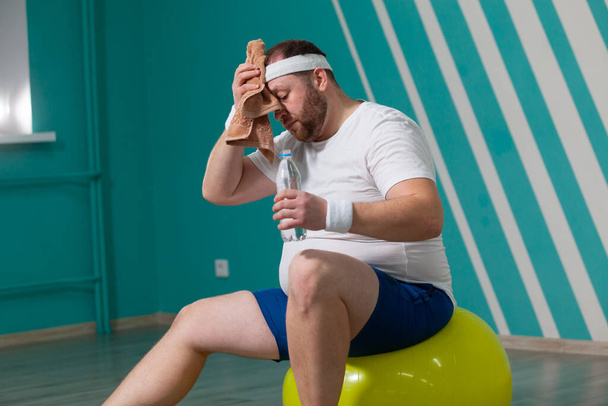 Overweight man is sitting on a fitness ball exhausted after a hard training in group fitness classes. Fat man uses a towel to wipe the sweat from his forehead - Photo, Image