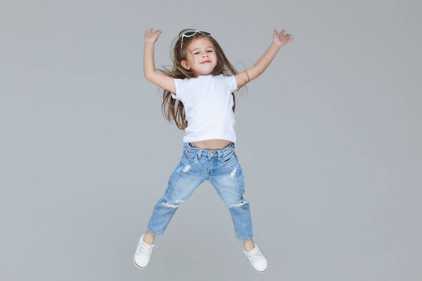 Kid girl preschooler in blue jeans, white t-shirt and sunglasses is jumping with raised hands isolated on grey background - 写真・画像