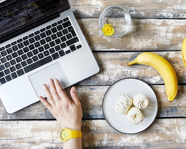 Жінка працює з дому і їсть банан і солодкий домашній Зефір або зефір з банана біля ноутбука на дерев'яному столі вид зверху. Час для перерви
 - Фото, зображення