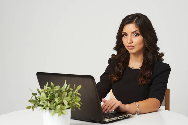 Een jong meisje met donker haar, een zwarte jurk, werkt thuis. Op het bureaublad staat een laptop en een pot bloemen. Geïsoleerde witte achtergrond meisje glimlachend. Werk geeft haar vreugde - Foto, afbeelding