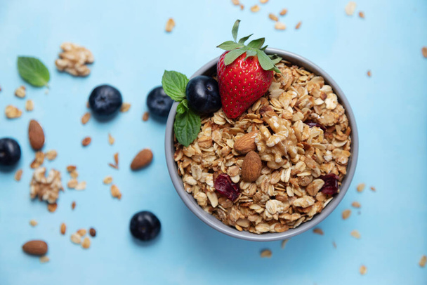 Bowl, cup with homemade granola or oatmeal granola with nuts and fresh strawberries and blueberries. Flakes and berries are scattered nearby - Fotó, kép
