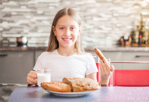 Una ragazza in età scolare è seduta in cucina. In una mano ha un bicchiere di latte, nell'altra un pezzo di pane fresco fatto in casa. Foto orizzontale
 - Foto, immagini