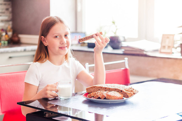 Dziewczyna w wieku szkolnym siedzi w kuchni. W jednej ręce ma szklankę mleka, w drugiej kawałek świeżego domowego chleba. Zdjęcie poziome - Zdjęcie, obraz