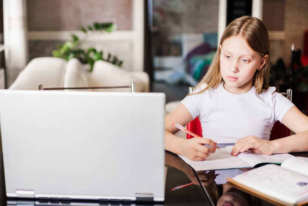 Okul çağındaki küçük kız evde okuyor. Bir kız dizüstü bilgisayar ekranından video dersi izler ve görevleri not defterine yazar. Yatay fotoğraf - Fotoğraf, Görsel