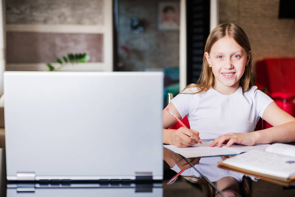 学校の年齢の少女は家で勉強している。女の子はノートパソコンのモニターでビデオレッスンを見て、ノートブックにタスクを書きます。女の子はカメラを見てください。横写真 - 写真・画像