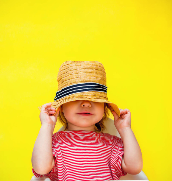 Little toddler girl hides her face with a straw hat. The child pulled a hat on his head on a yellow background. Copy space and place for text. - Φωτογραφία, εικόνα