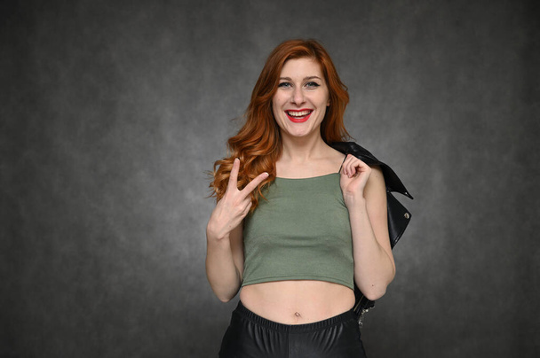 Pretty model actress posing with different emotions on a gray background in the studio. Portrait of a young Caucasian woman with long red hair in a green T-shirt and with a black jacket in her hands. - Φωτογραφία, εικόνα