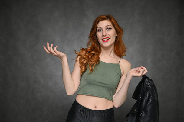 Pretty model actress posing with different emotions on a gray background in the studio. Portrait of a young Caucasian woman with long red hair in a green T-shirt and with a black jacket in her hands. - Фото, изображение
