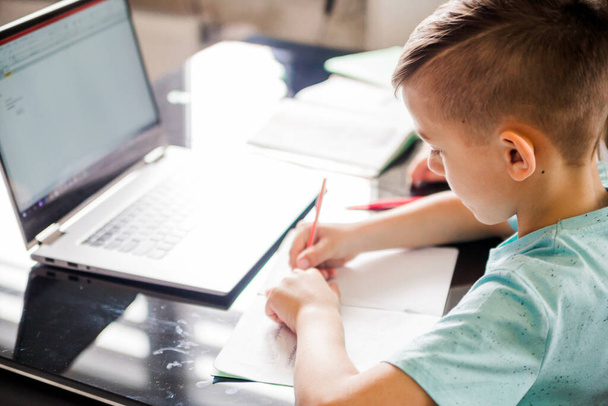 El chico está estudiando en casa. Se sienta en una mesa con libros de texto, un portátil está abierto delante de un niño. Fotografía horizontal
 - Foto, imagen