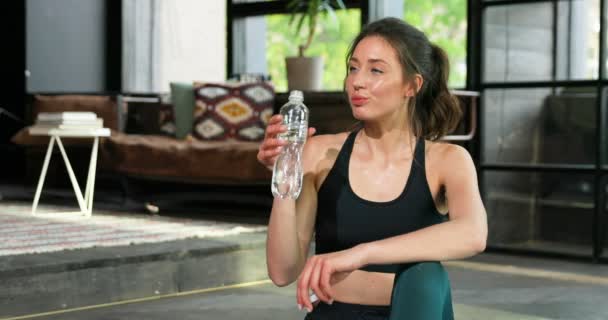 Sporty beautiful young Caucasian woman drinking water and resting after training at home. Sweaty pretty female sipping cold drink after working out in living room on quarantine Sport on self-isolation - Imágenes, Vídeo