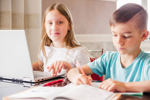 Boy and girl, brother and sister study at home. A girl is watching a video lesson or an online conference, the boy is doing exercises in a notebook from a textbook. Girl looking at the camera. Horizontal photo - 写真・画像