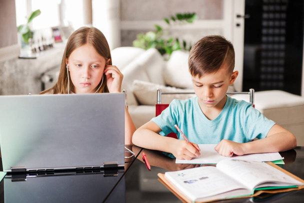 Chłopiec i dziewczynka, brat i siostra uczą się w domu. Dziewczyna ogląda lekcje wideo lub konferencję online, chłopak robi ćwiczenia w notatniku z podręcznika. Zdjęcie poziome - Zdjęcie, obraz