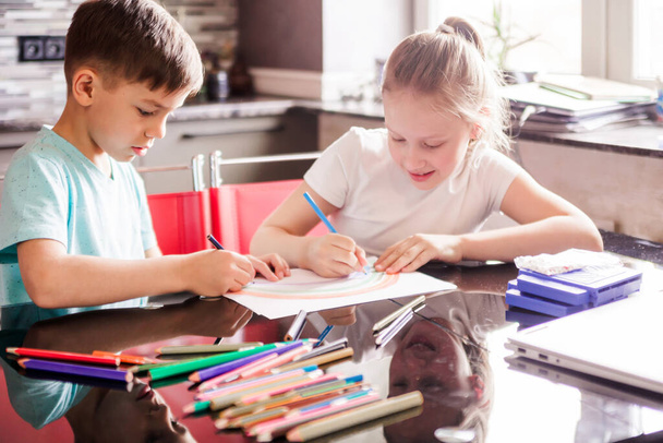 Chłopiec i dziewczyna, brat i siostra narysować tęczę z kredkami razem, siedzi przy stole. Zdjęcie poziome - Zdjęcie, obraz
