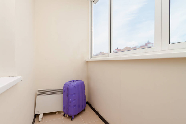 Maleta de plástico violeta en un rincón vacío de la habitación del balcón con ventana acristalada y vista al cielo azul nublado. Quédese en casa, no viaje o simplemente llegue o deje el concepto
 - Foto, imagen
