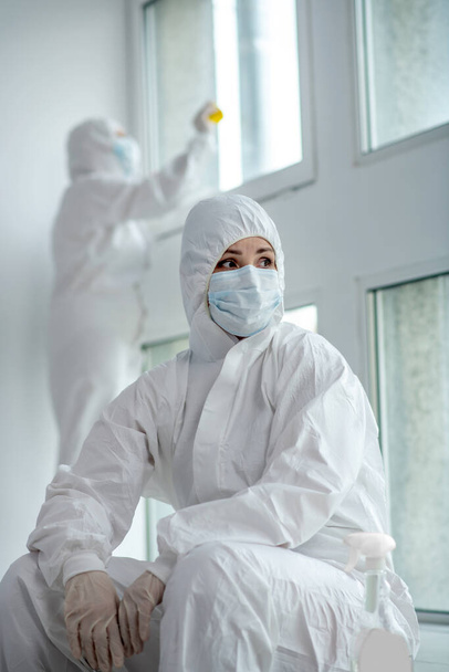 Medical worker in protective clothing and medical mask sitting next to the window, her collegue desinfecting panes - Photo, Image
