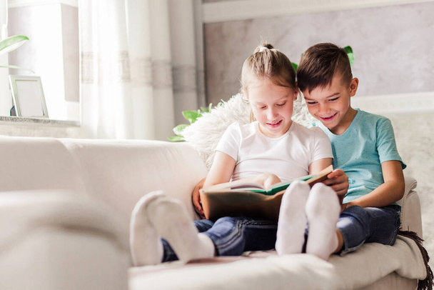 Boy and girl, brother and sister read a book together while sitting on a sofa at home. Horizontal photo - Фото, изображение